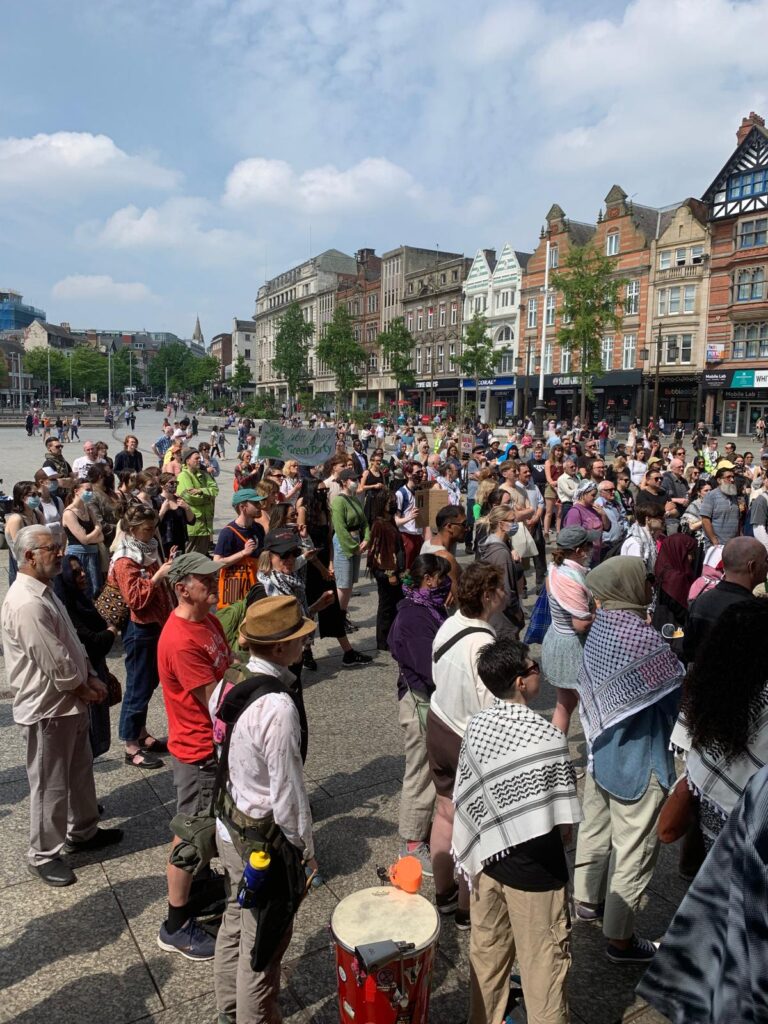 A photograph of a large group of people attending a pro-Palestine demonstration in Nottingham's Old Market Square. They are facing a speaker off-screen to the right of the photo. The Nottingham Green Party banner is visible amongst the group.