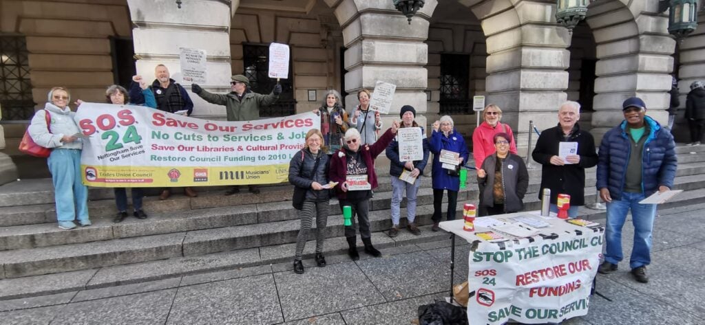 A photograph of a group of people outside the Council House in Nottingham facing the camera holding signs and banners supporting the Save Our Services 24 campaign.